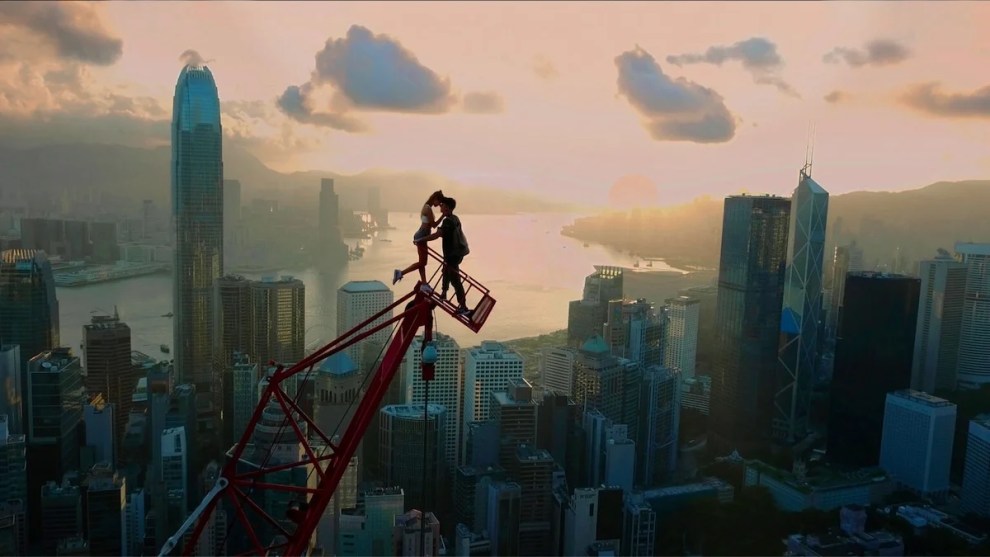 A man and a woman share an intimate moment atop a crane, high above a cityscape with the sun lighting up the sky in the background from the horizon.